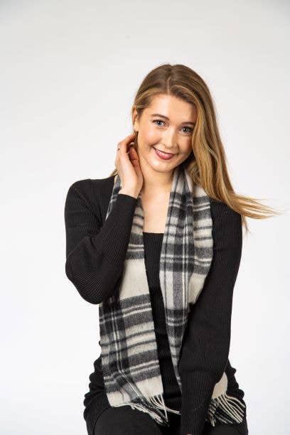 A woman with long blonde hair smiles and touches her neck against a white background, wearing a black outfit and the Stewart Grey Dress Lambswool Scarf.