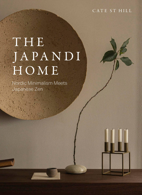 A minimalist interior inspired by Japandi style, featuring Japandi Home by Cate St Hil: Nordic Minimalism Meets Japanese Zen, a round textured wall piece, vase with branch, stacked dishes, and a modern candle holder on wood.
