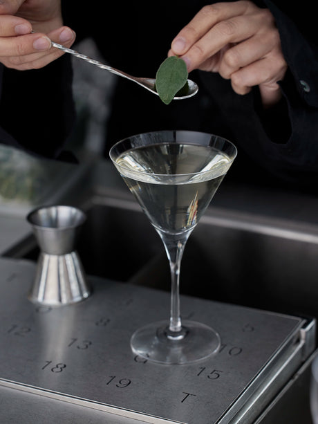 A person elegantly garnishing an Orrefors: More Martini Glass 6.4oz with a leaf, using a spoon.