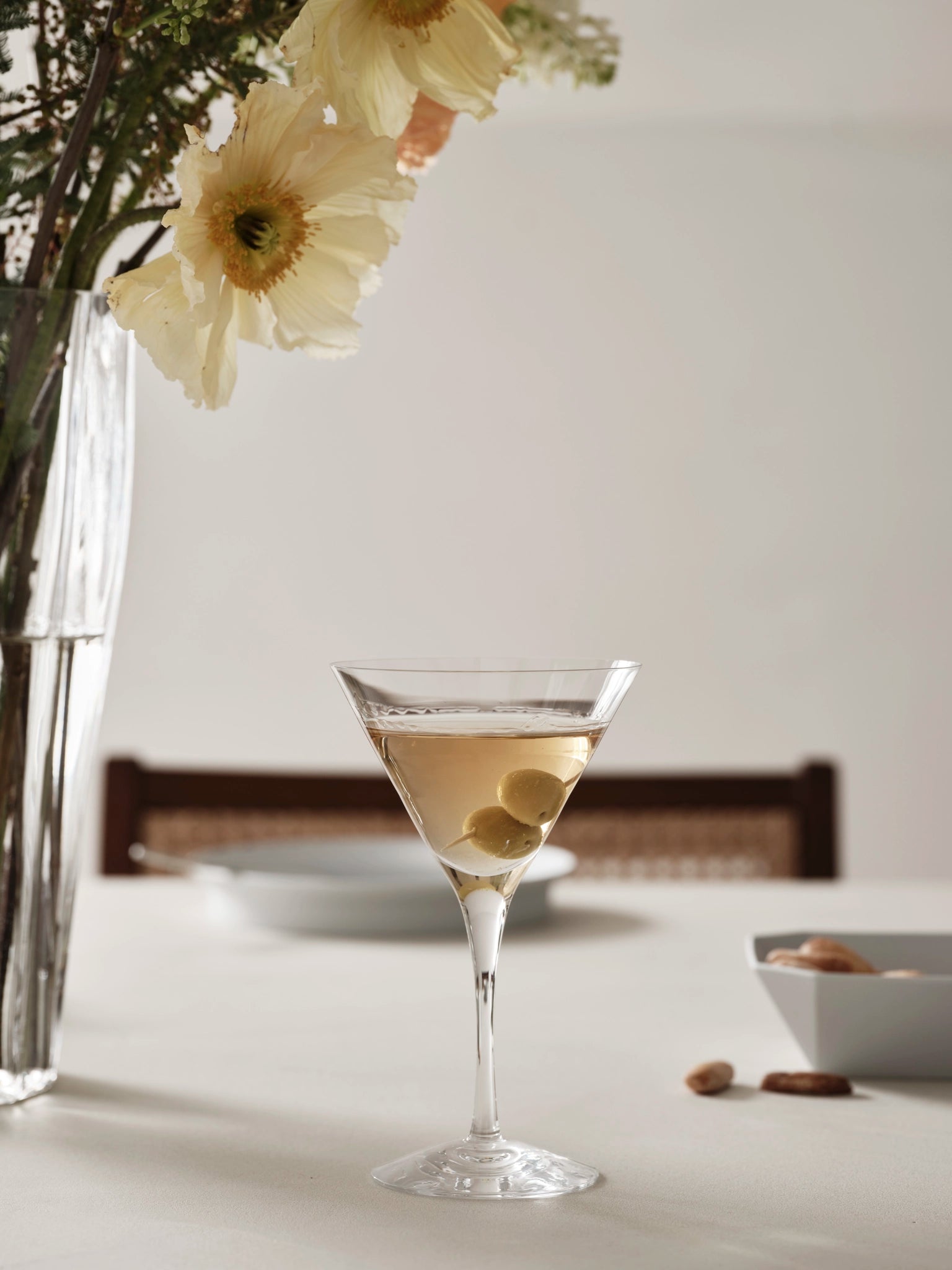 An Orrefors: More Martini Glass 6.4oz from the 2-pack, filled with olives, sits beside a large flower vase and a bowl of nuts, showcasing Orrefors elegance.