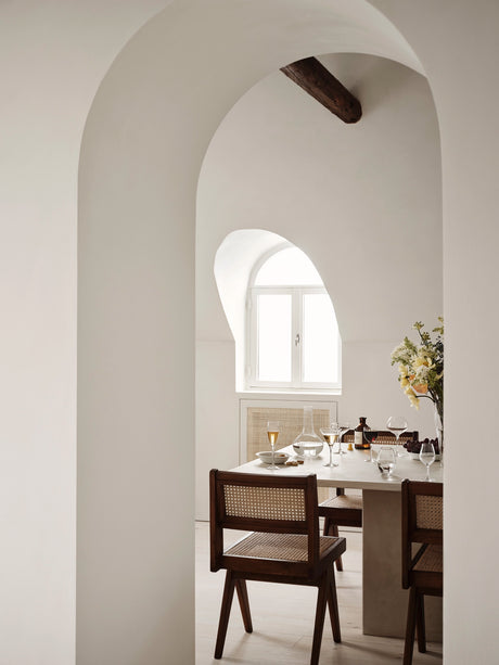 An arched doorway opens to a dining area with a light table and four chairs. The table is elegantly set with Orrefors More Martini Glasses, bottles, and a flower vase beneath a bright window framed by wooden beams.