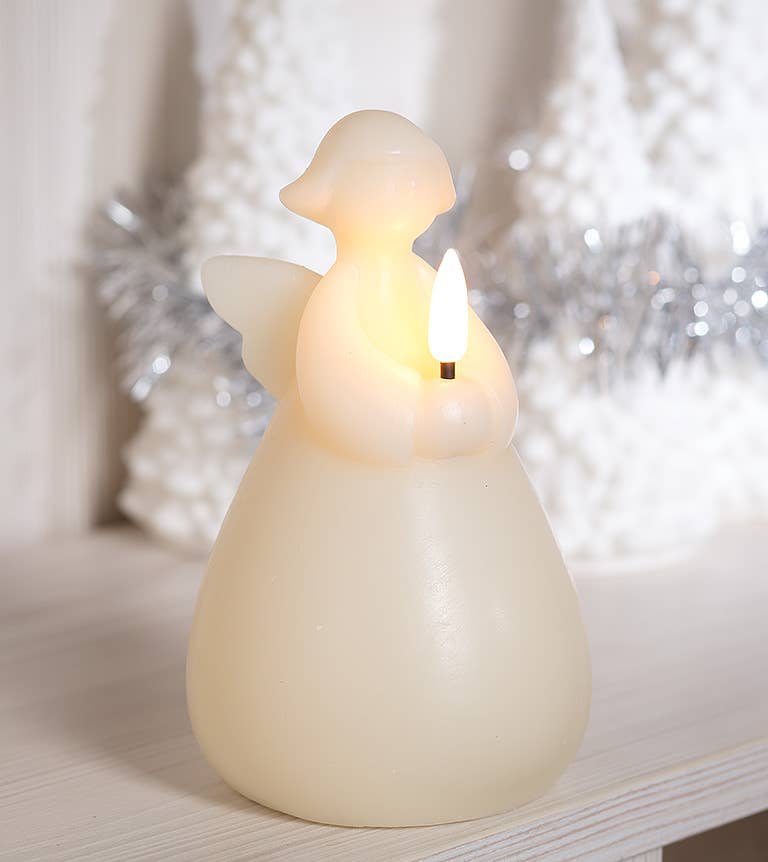 The Candle: Angel LED - Ivory 6" casts a warm ambient glow as it rests on the table, encircled by white decorative trees and silver tinsel, creating a magical atmosphere with its perfectly timed 6-hour timer.