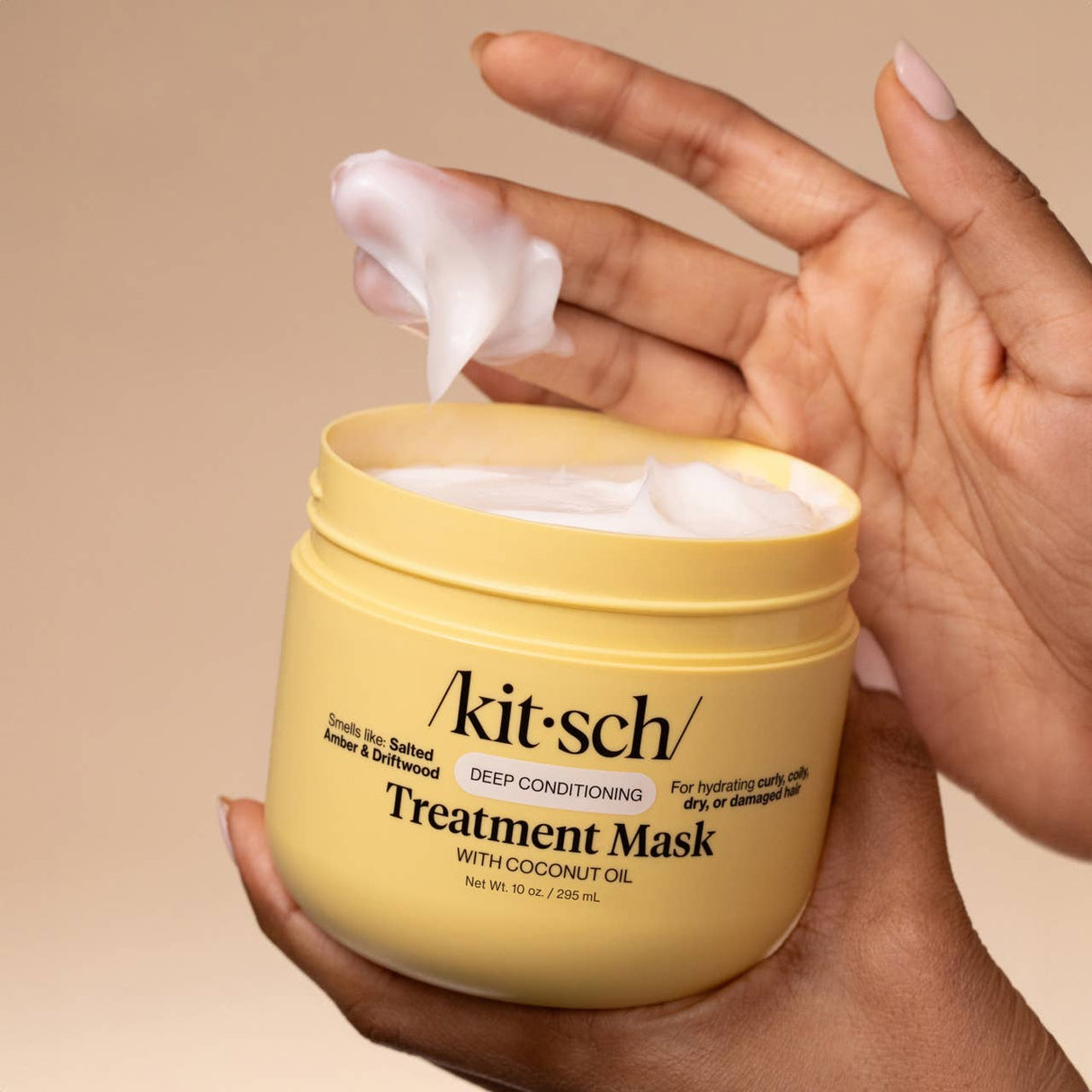 A hand scoops white cream from an open yellow jar labeled Hair: Moisturizing Coconut Oil Deep Conditioning Treatment Mask 10 oz by Kitsch.