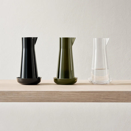 The Orrefors: Informal Carafe with Bowl (Olive), with its mouth-blown elegance, is displayed on a light wooden shelf, its timeless design standing out against the plain wall.