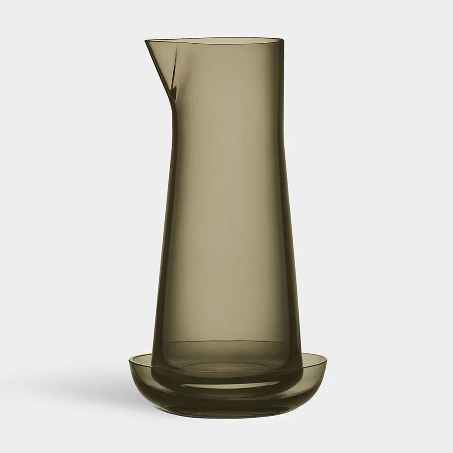 The Orrefors Informal Carafe with Bowl (Olive) features a tall, translucent, olive-green mouth-blown glass design with a spout, displayed on a matching round glass base against a plain light background.