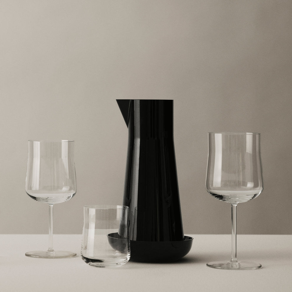 The Orrefors: Carafe with Bowl, in black, sits empty on a white surface between a short glass and two wine glasses, all against a neutral background.