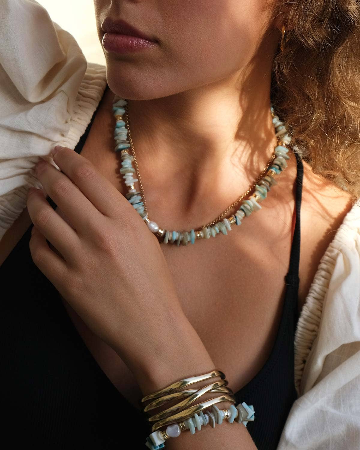 Wearing the A&C Oslo Blue Shell Necklace, this person complements their turquoise and gold outfit. Curly hair falls over a white blouse with black accents as they softly touch the semi-precious stones that finalize their look.