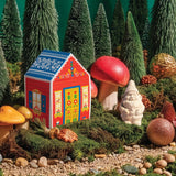 A colorful model house, white gnome, red mushrooms, and ornaments arranged among green trees, moss, and Swedish Pine in a forest setting.