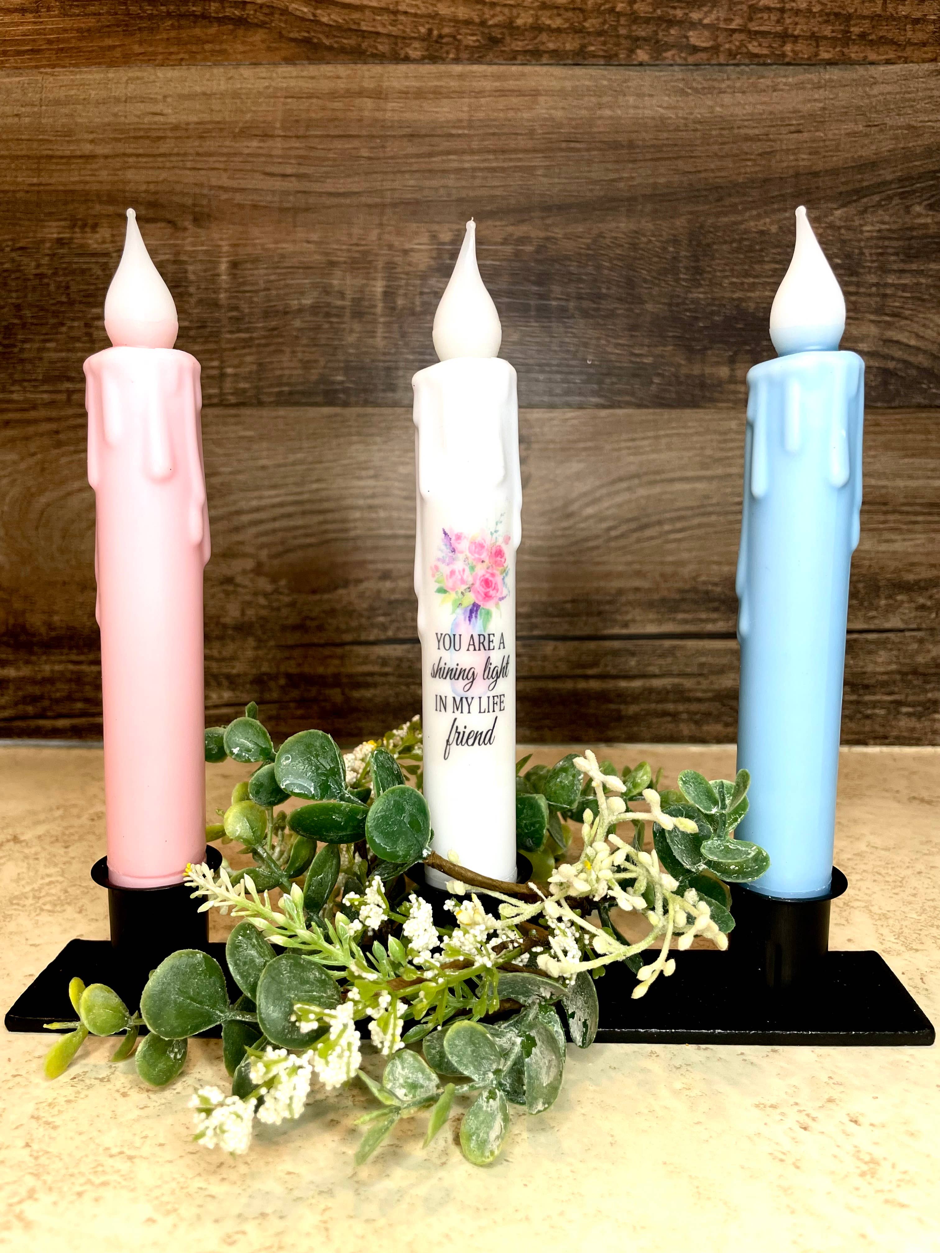 The Candle: Friend Sentiment LED Battery Operated with Timer Taper Candles includes three decorative candles—pink, quote-adorned white, and blue—on a black stand with green and white foliage. They add charm against a wood-textured wall without the worry of open flames.