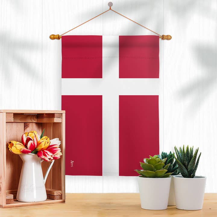 The Denmark Garden Flag – Danish Nationality Home Decor Flag hangs on a white wall above a small table with potted succulents and a wooden box holding a white jug with colorful tulips.