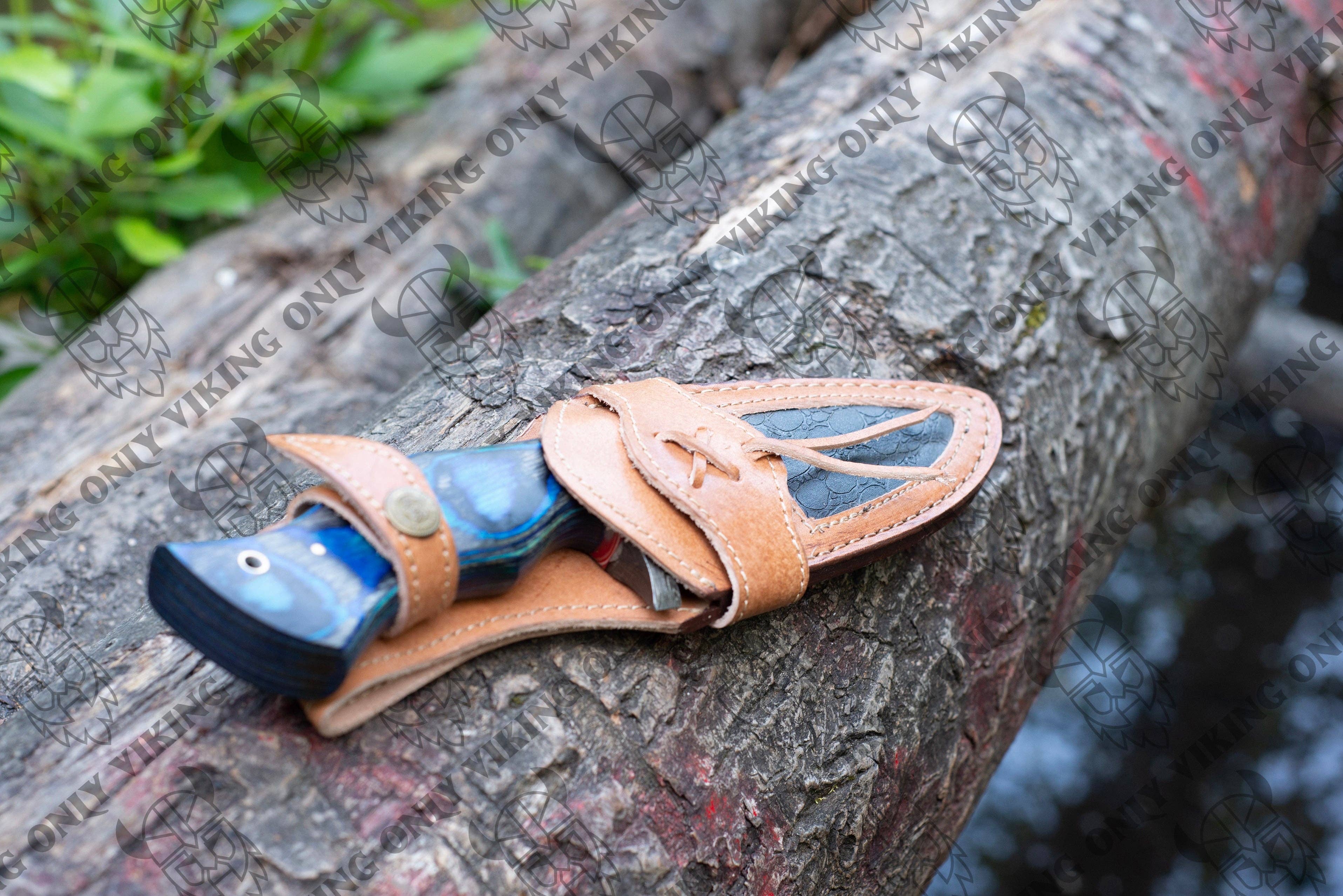 The Premium Quality Damascus Utility Knife, featuring a blue and black handle and crafted from hand-forged Damascus steel, rests in a tan leather sheath on a tree trunk amidst lush greenery.