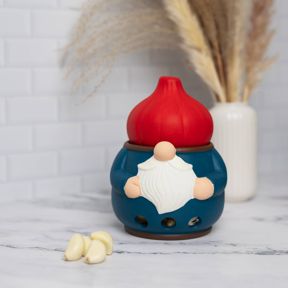 Ceramic gnome garlic keeper with a red hat on a marble counter, garlic cloves beside it, and dried grass in a white vase in the background.