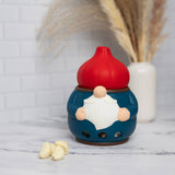 Ceramic gnome garlic keeper with a red hat on a marble counter, garlic cloves beside it, and dried grass in a white vase in the background.