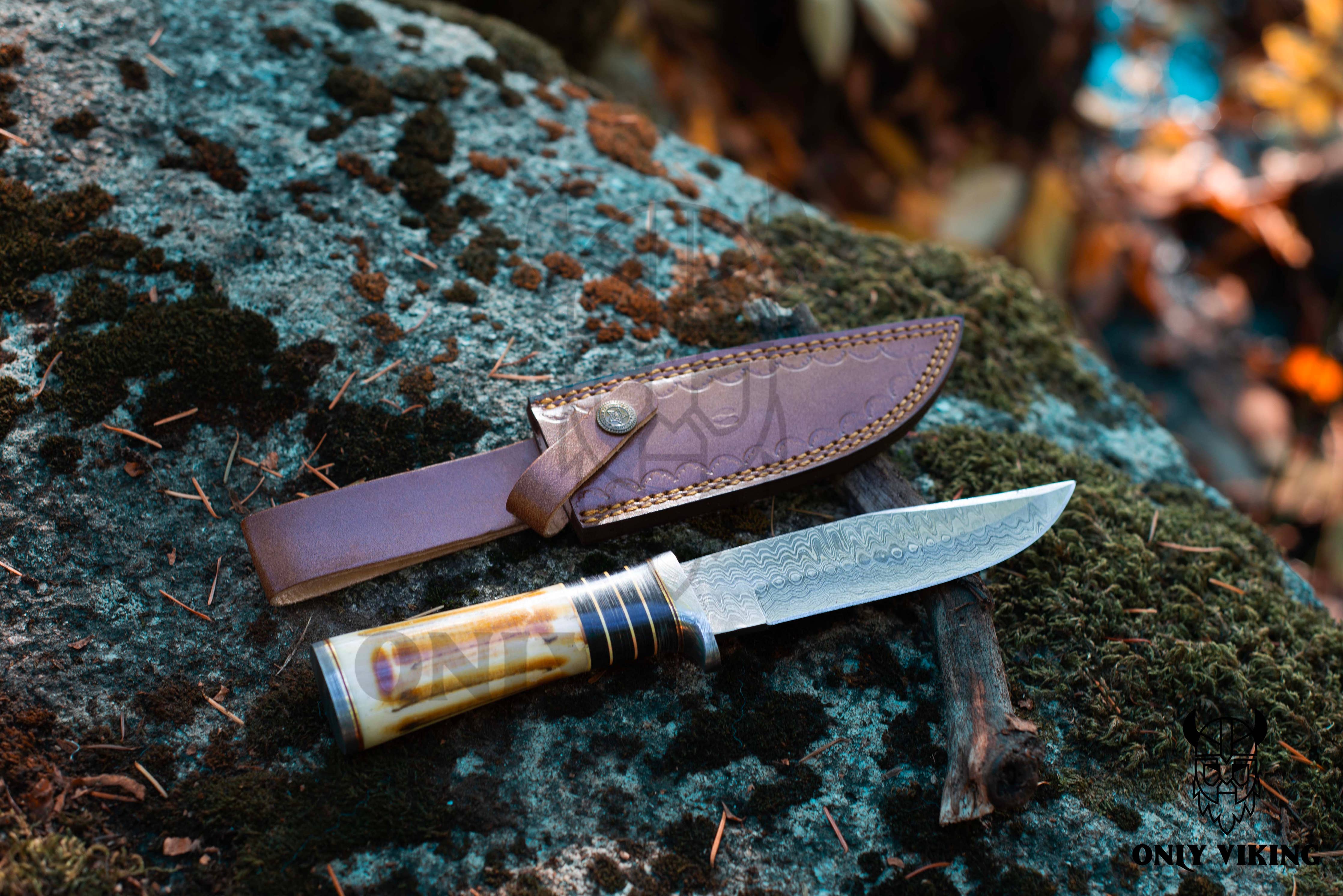 A Knife: Damascus Hunting Knife with Locking Leather Sheath rests on a mossy rock, showcasing its patterned blade and handle, ready for outdoor adventures.