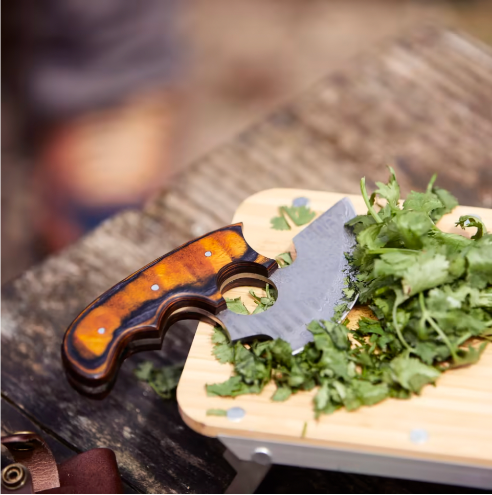 The Alaskan Ulu Style Blade knife with a wooden handle sits on a cutting board beside freshly chopped cilantro on an outdoor wooden surface.