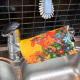 The Light of Grytnäs Dala Horse dish cloth with a colorful floral design hangs over a silver kitchen faucet, alongside an eco-friendly white scrub brush. The black tiled backsplash and stainless steel sink highlight its super absorbency for efficient kitchen cleaning.