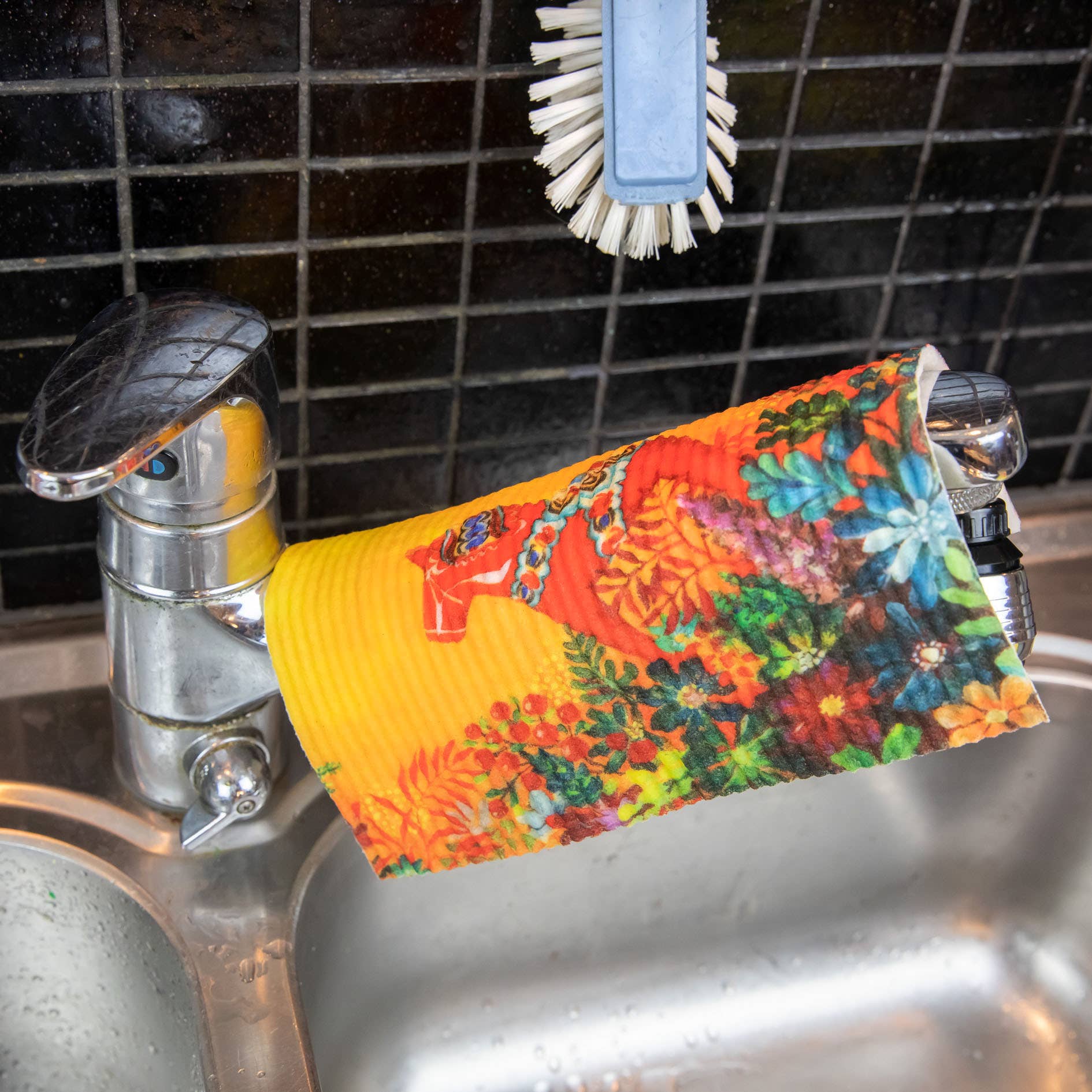 The Light of Grytnäs Dala Horse dish cloth with a colorful floral design hangs over a silver kitchen faucet, alongside an eco-friendly white scrub brush. The black tiled backsplash and stainless steel sink highlight its super absorbency for efficient kitchen cleaning.