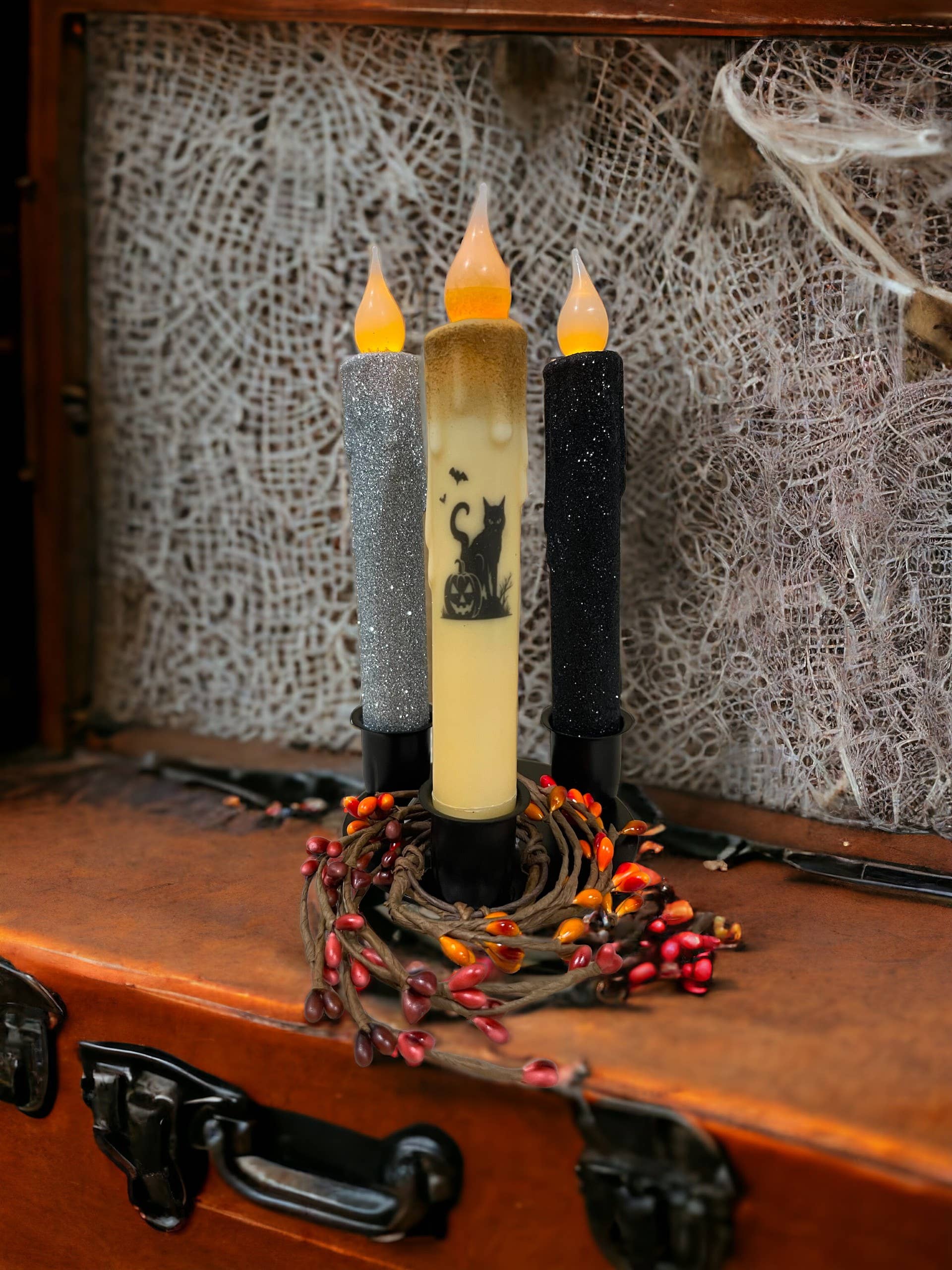 Three LED candles—silver, cream with a cat and pumpkin design, and black—are displayed on a Round Triple Wrought Iron Taper Holder by Lancaster Blacksmith Products in a Halloween-themed setting, surrounded by a wreath of red berries.