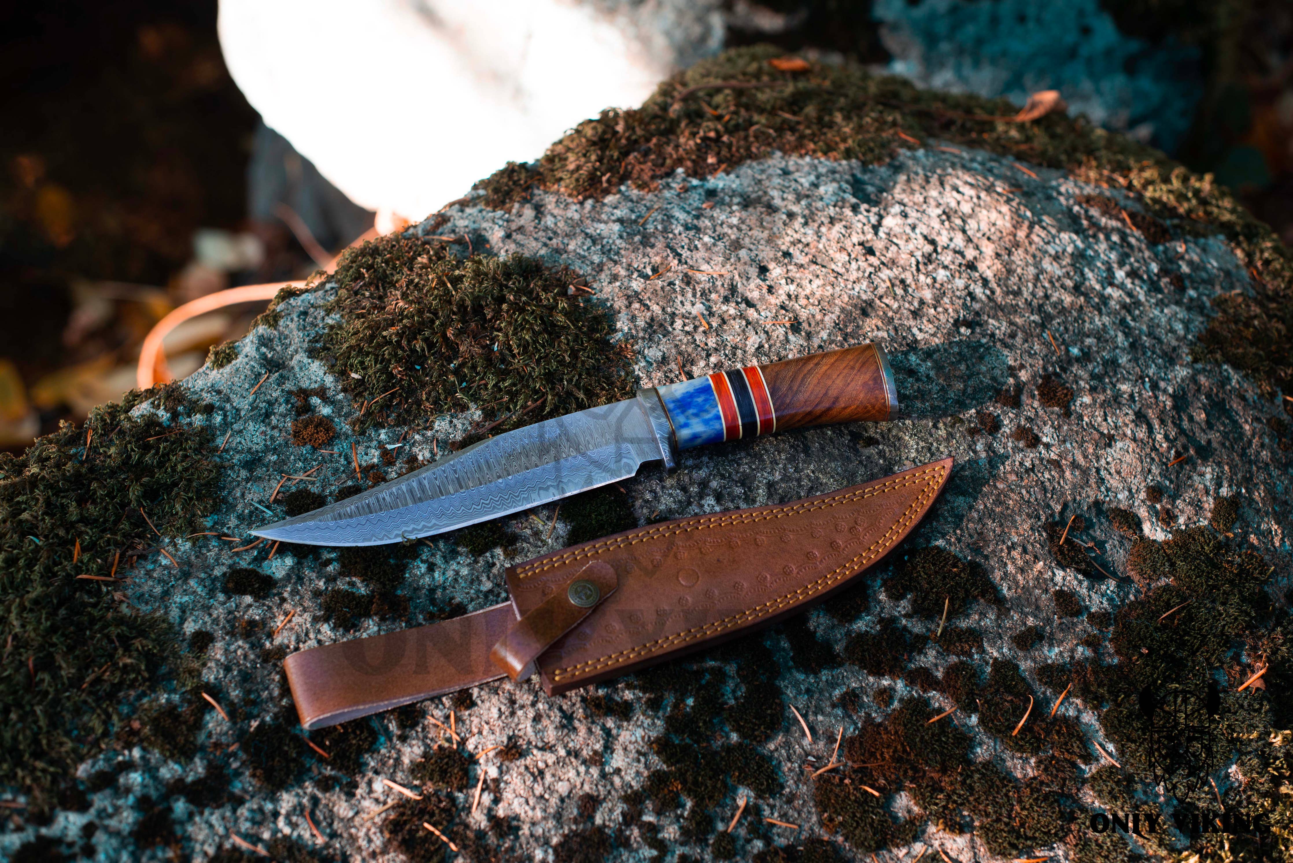 The Damascus Steel Hunting Knife, featuring a decorative folding design with a striped handle and a leather sheath, rests on the moss-covered rock. Its blade glistens with the distinctive pattern unique to Damascus steel.