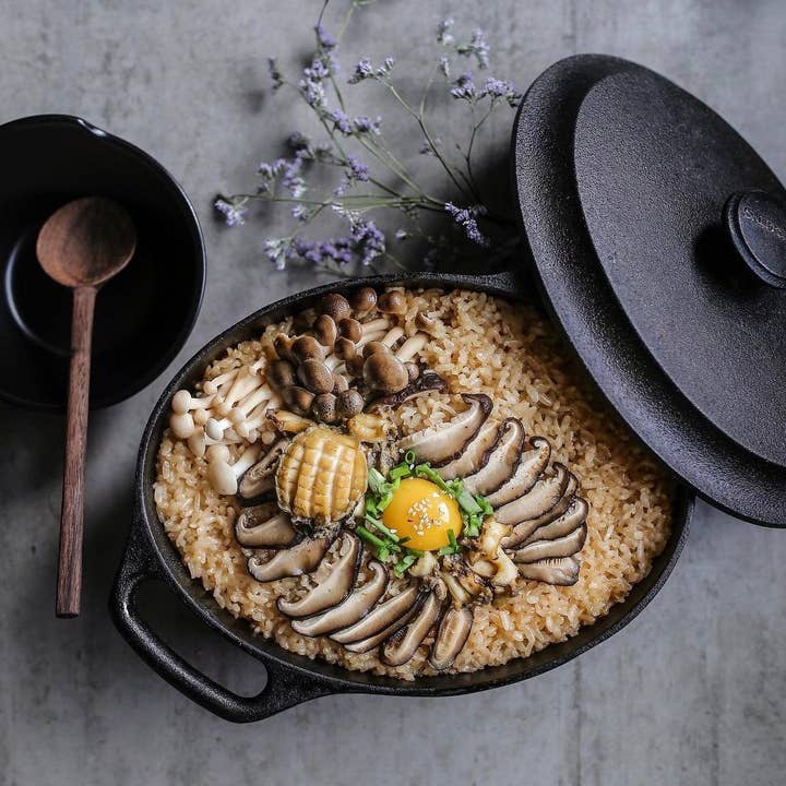 The Skeppshult Oval Dutch Oven With Cast Iron Lid 6.3 qt is filled with rice, mushrooms, abalone, a raw egg yolk, and green onions, served with a bowl and wooden spoon on the side.