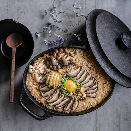 The Skeppshult Oval Dutch Oven With Cast Iron Lid 6.3 qt is filled with rice, mushrooms, abalone, a raw egg yolk, and green onions, served with a bowl and wooden spoon on the side.