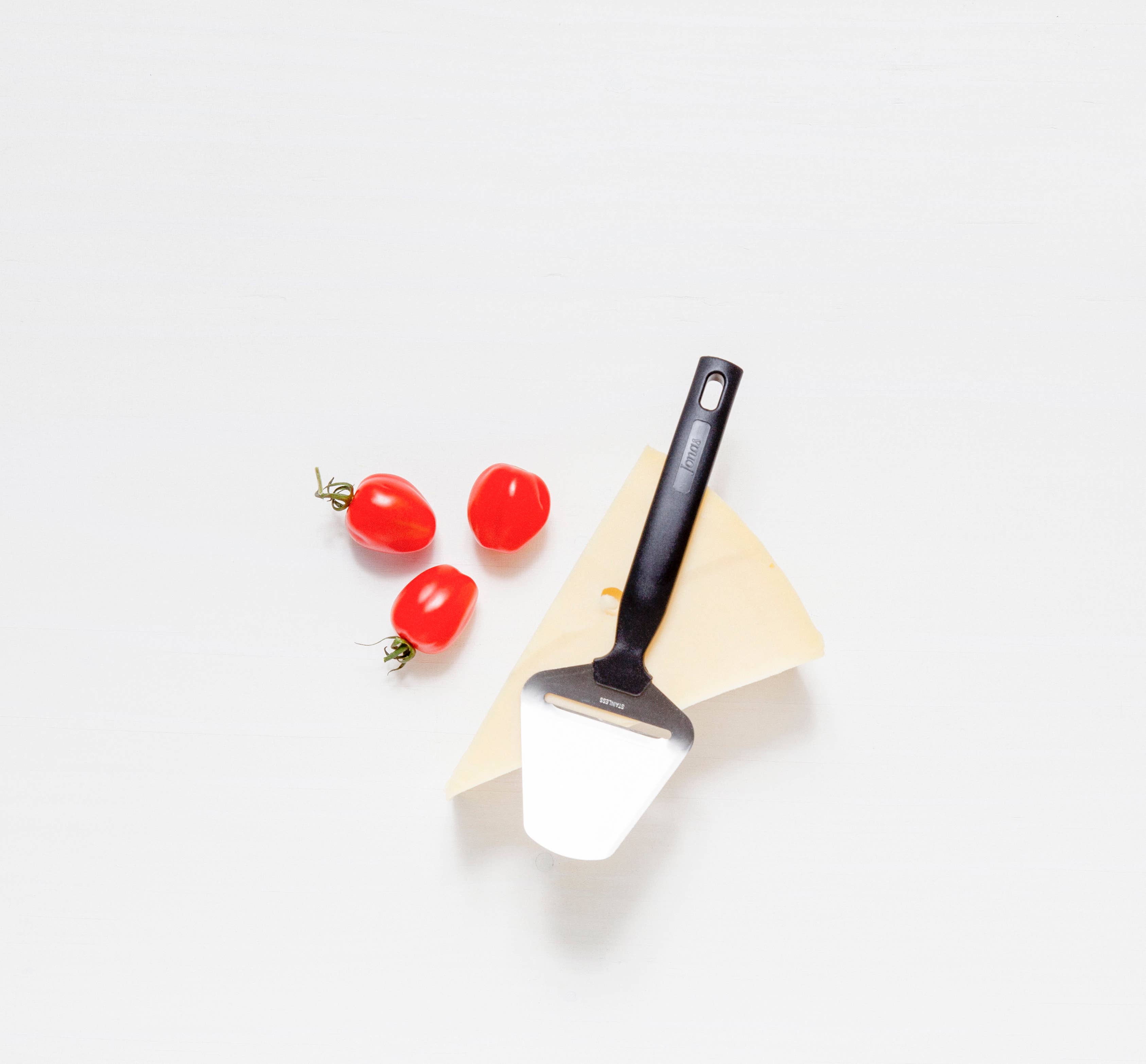 A Swedish Cheese Plane for Hard Cheese with a stainless steel blade lies on a hard cheese wedge, alongside three ripe red tomatoes on a white surface.