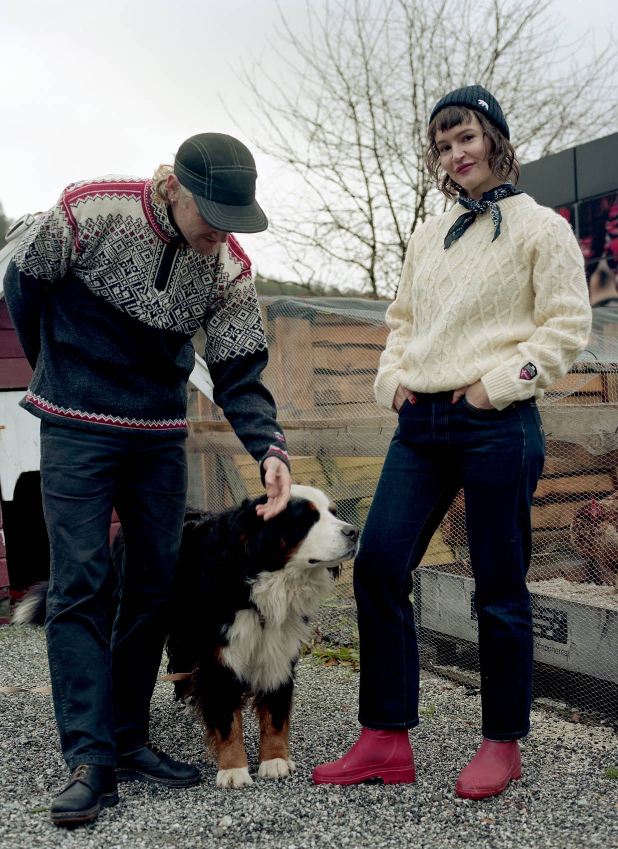 Two people stand outside on gravel with a large black, white, and brown dog; one wears the Norlender Knitwear Narvik Ski Sweater (312) in Charcoal and pets the dog while the other stands nearby with hands in pockets.
