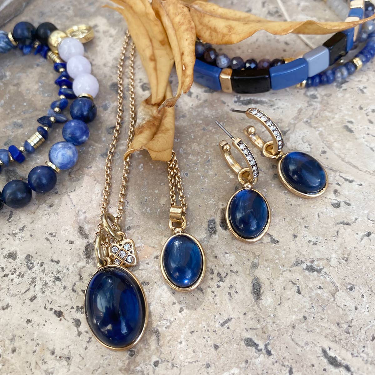 The Sophistication Collection includes a blue and gold jewelry set with matching necklaces, earrings, and bracelets elegantly displayed on a stone surface with a dried leaf. Featuring the A&C Oslo Gold Plated Glass and Crystal Pendant Necklace, each piece exudes timeless charm.