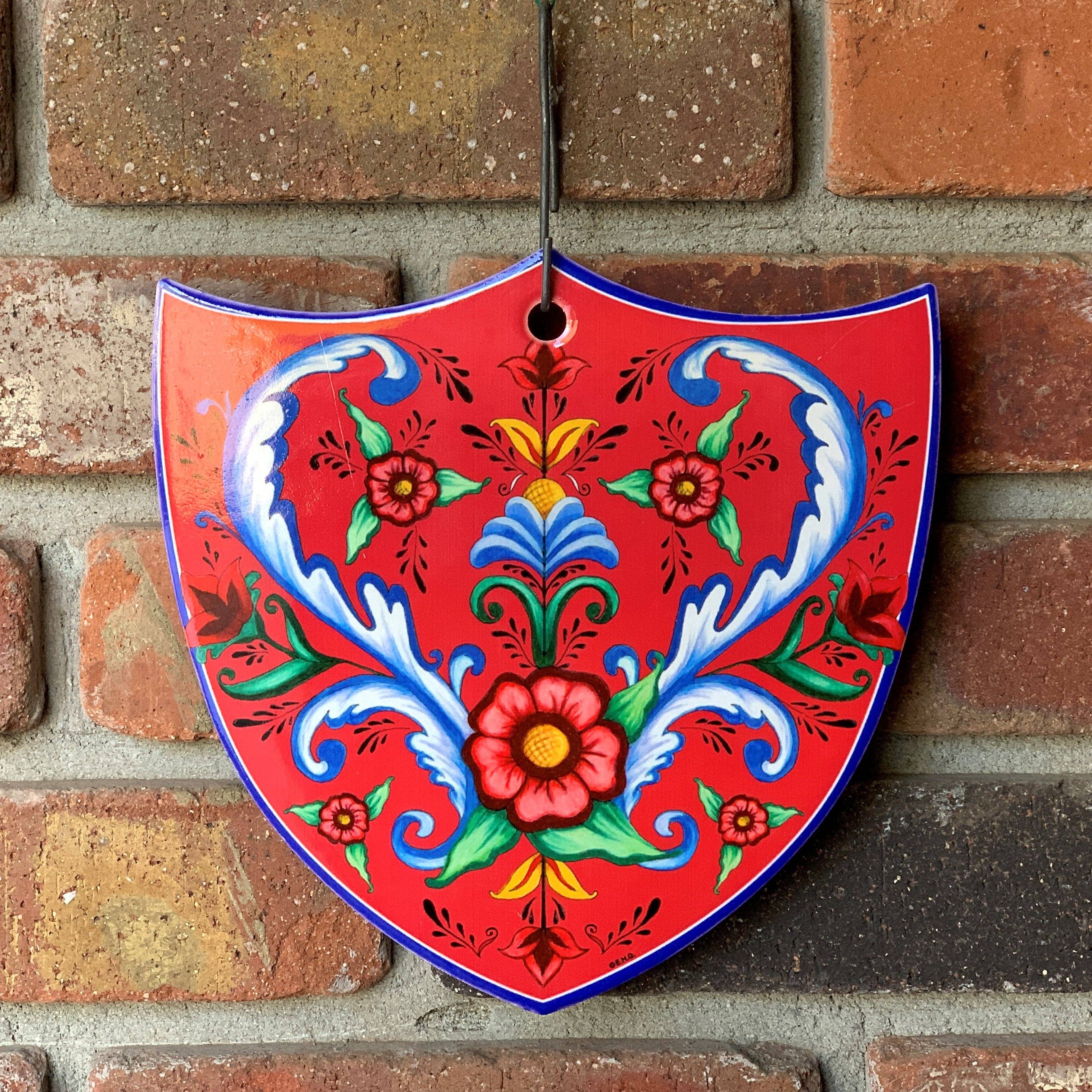 The "Norwegian Rosemaling - Flowers Decoration Shield Tile," a red ceramic wall hanging with blue trim and multi-colored floral and leaf patterns, is displayed on a brick wall. The tile measures 7.75" high and is inspired by traditional Norwegian design.