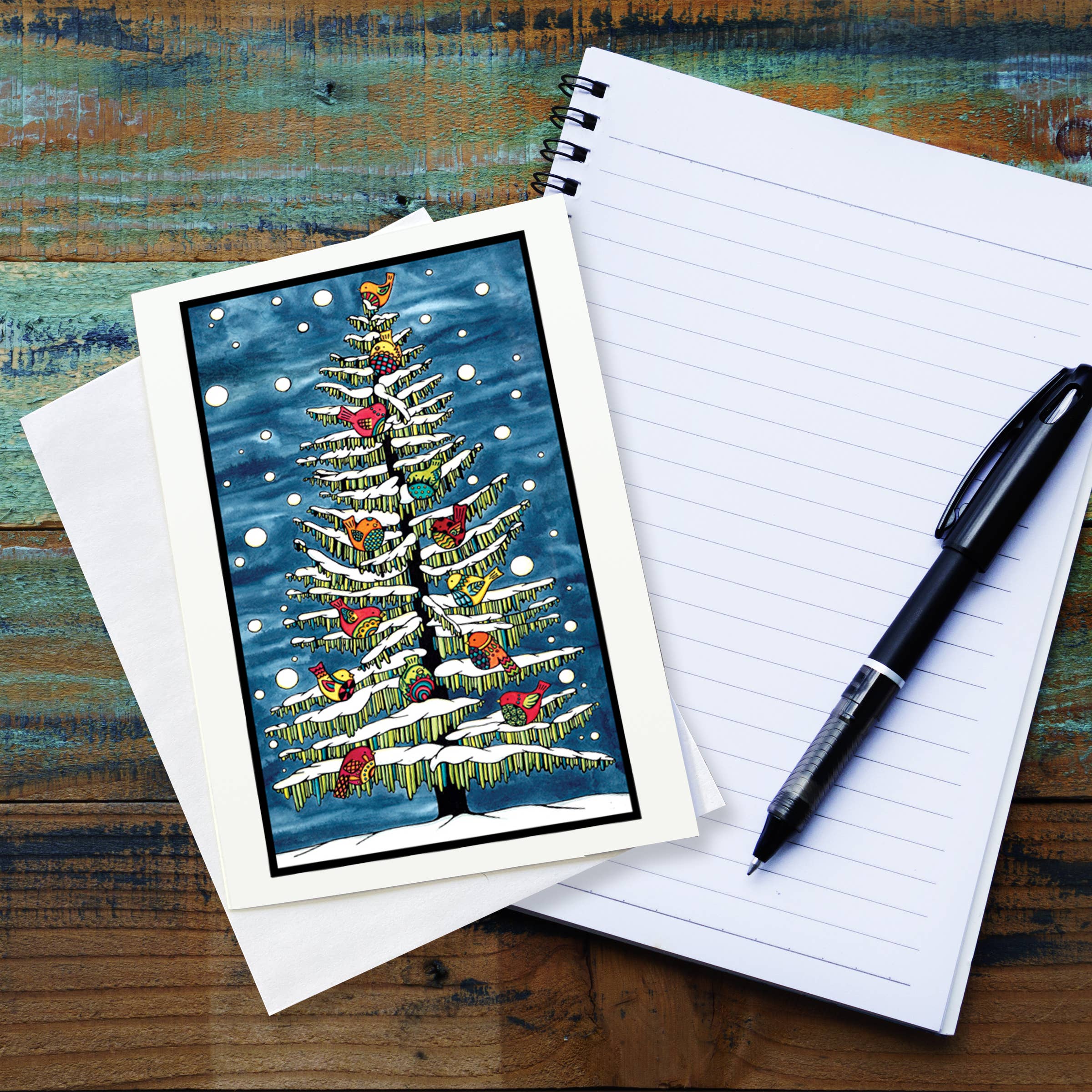 The Card: Holiday Birds in Snow Greeting Card by Sarah Angst (5x7, with envelope) sits atop a blank notepad and black pen on a wooden surface. The card is blank inside for your personal message.
