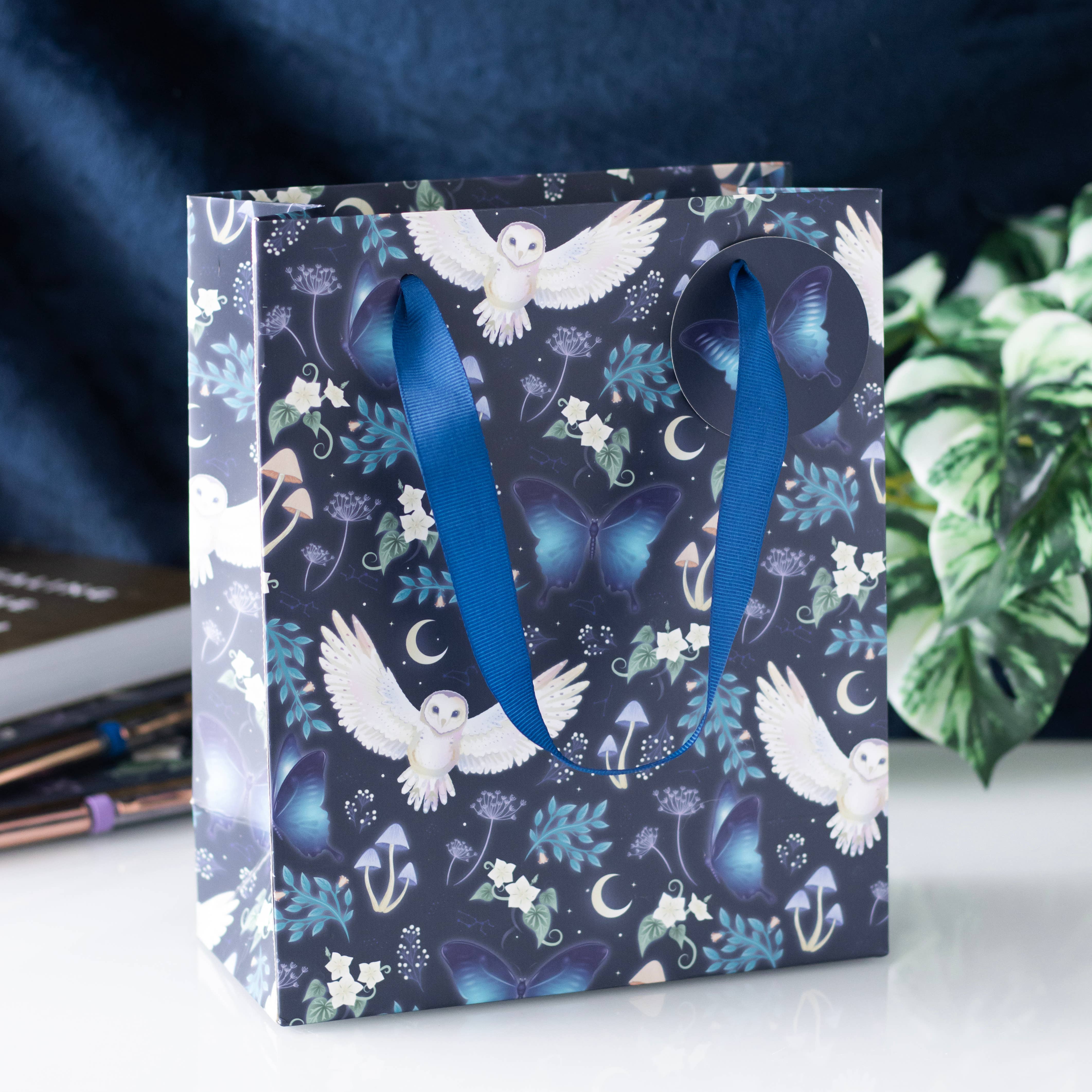 The Midnight Forest Owl Print Medium Gift Bag, featuring white owls, blue butterflies, anchors, moons, stars, green flowers, and a blue ribbon handle, sits on a white surface.