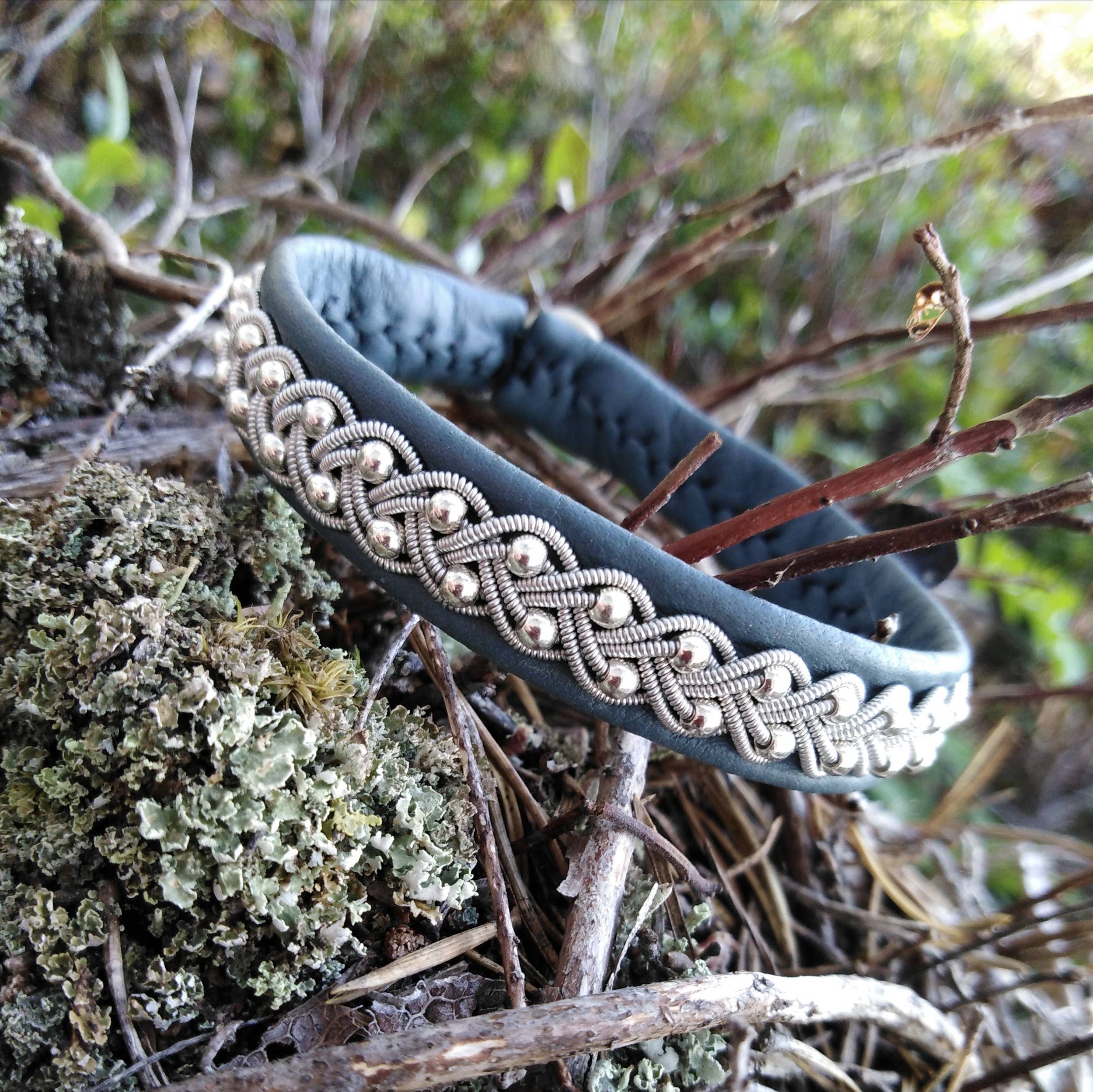 The Sami Lapland Bracelet (#39), crafted from reindeer hide with pewter, silver, and gold beads in black (17-18 cm), is displayed resting on mossy branches and twigs outdoors.