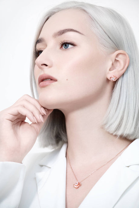 A woman with light skin and platinum blonde hair wears a white outfit and the Ice Dance Rose Gold/Sterling Silver Necklace with Brilliant Cut Cubic Zirconia, gazing thoughtfully to the side with her hand near her chin.