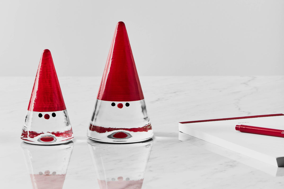 Two hand-painted glass gnome figurines with red pointed hats and minimalist facial features, standing on a marble surface next to a notebook and pen.