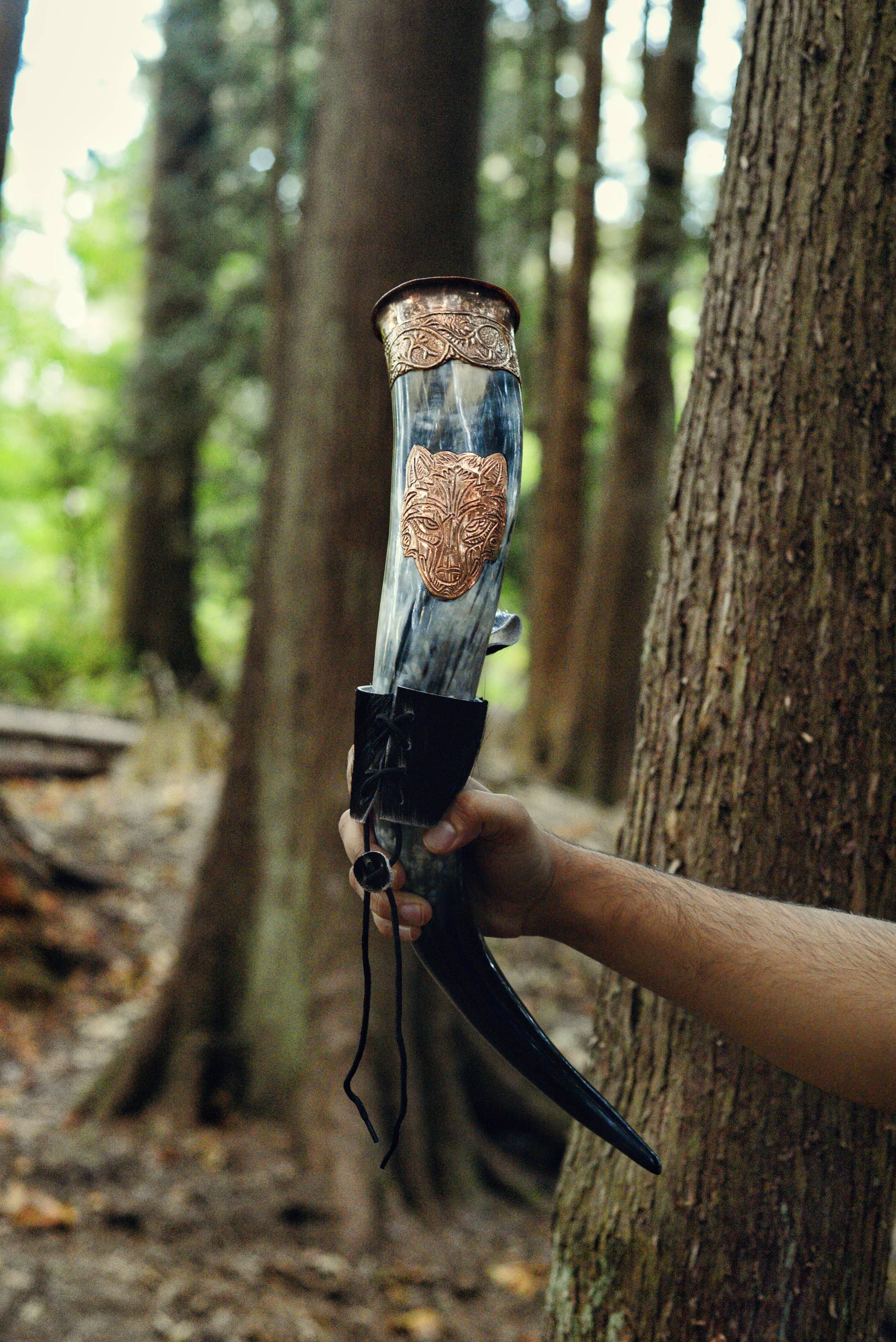A person holds the Drinking Horn: Authentic Viking Wedding Gift, crafted from real ox horn with metal accents and a wolf emblem, set against a forest—perfect for history lovers.
