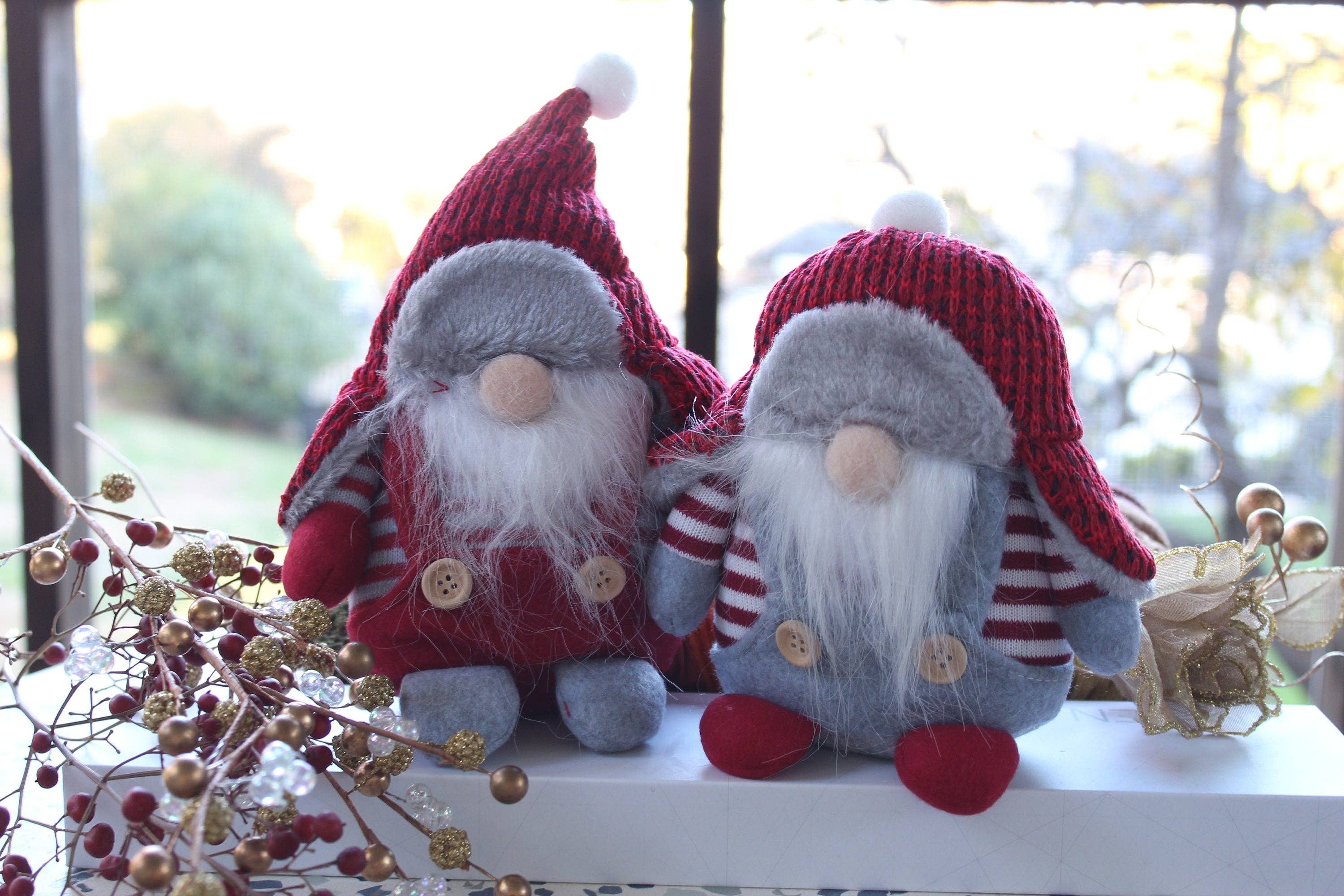 Two Winter Gnomes, named Aldrich and Bernard, inspired by Scandinavian folklore with white beards and red hats, sit on a ledge surrounded by festive decorations and berries.