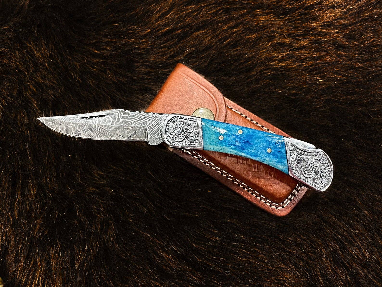 The Knife: Dyed Blue Bone 6.5 Damascus Steel features ornate silver detailing and a blue handle, presented on a brown leather sheath against a dark background—an elegant choice for collectors.