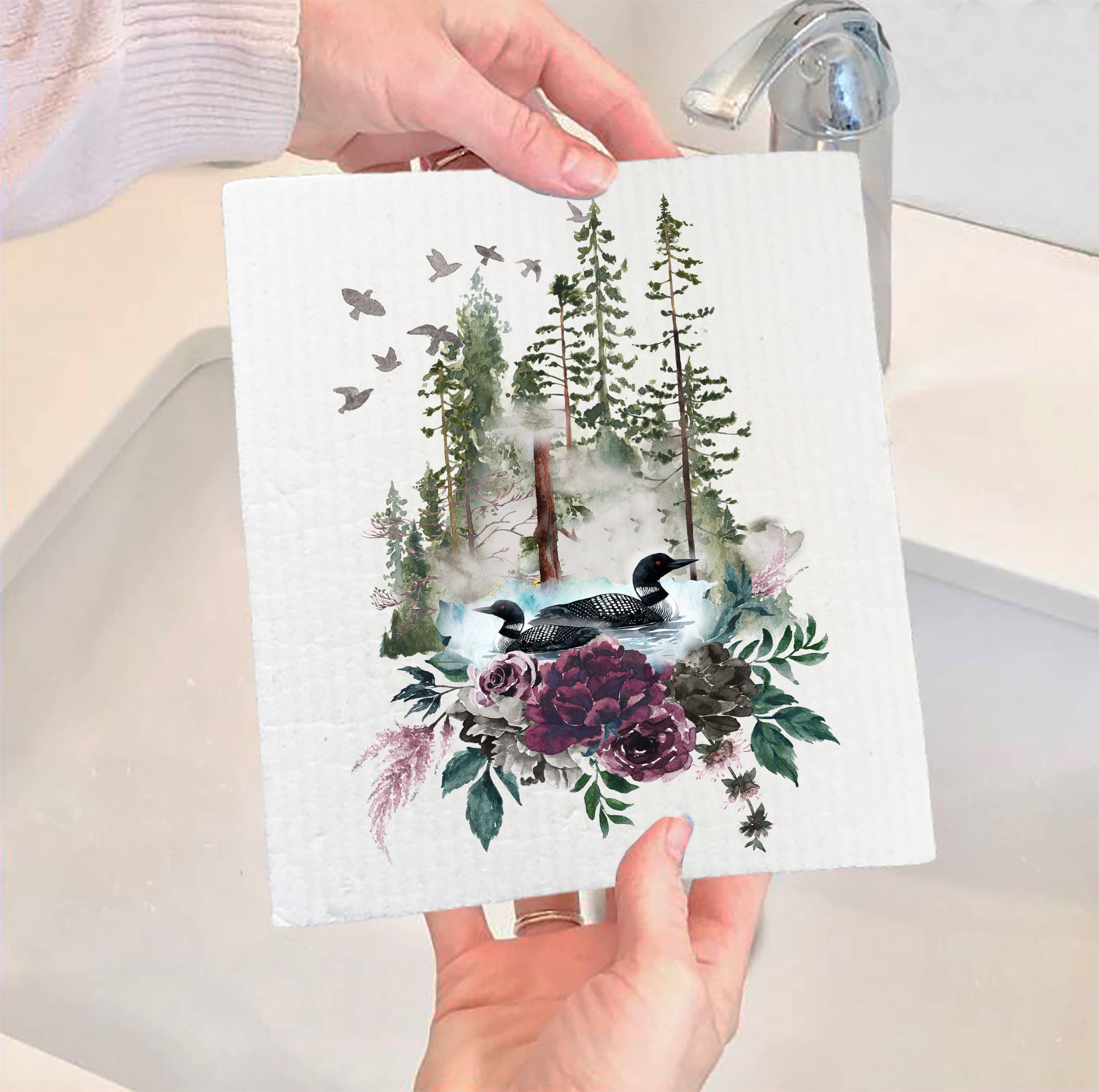 Hands holding a Swedish dishcloth near a sink, featuring artwork of loons, lakes, and mountains. The eco-friendly Swedish Dish Cloth: Loons Lake Mountains Wildlife is biodegradable, offering both charm and sustainability for your kitchen.