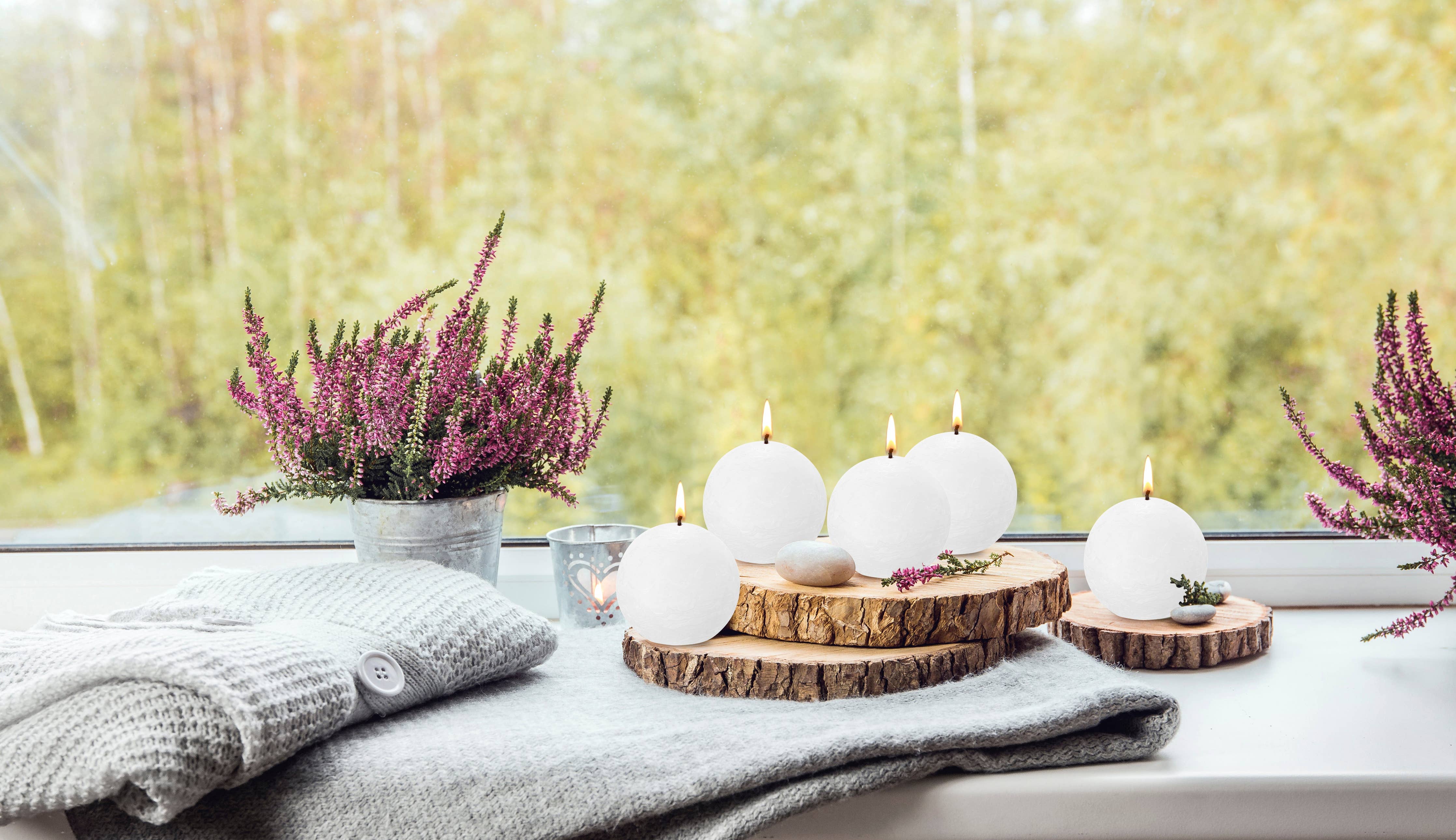 A cozy window scene showcases eco-friendly Blossom Pink Rustic 3" Ball Candles made from plant-based wax on wooden slabs, alongside a pot of purple flowers, a folded gray sweater, and a soft blanket, all set against a blurred outdoor background.