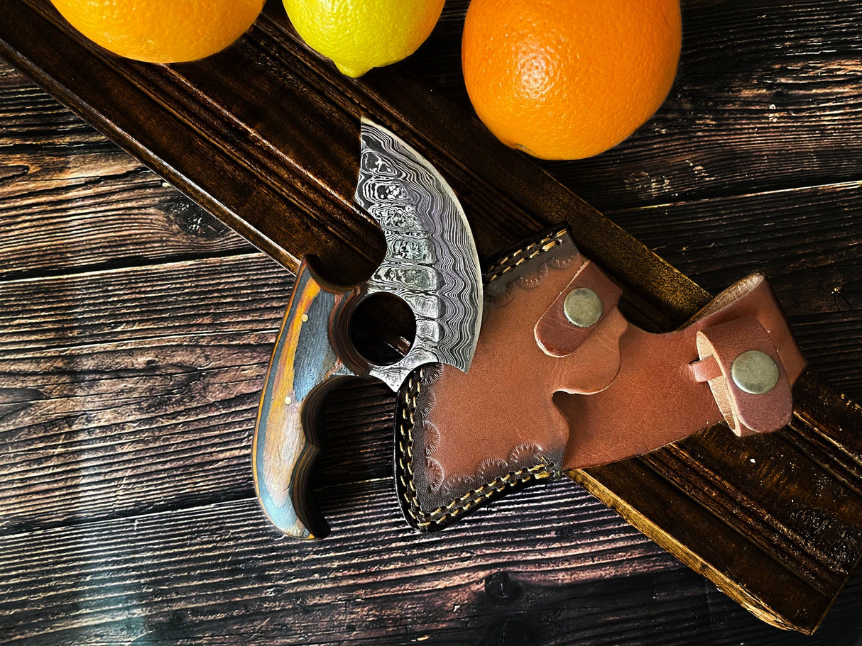 The Knife: Alaskan Ulu Style Blade, featuring a curved Damascus steel blade, ergonomic wooden handle, and leather sheath, rests on a wooden surface next to oranges and a lemon.