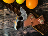 The Knife: Alaskan Ulu Style Blade, featuring a curved Damascus steel blade, ergonomic wooden handle, and leather sheath, rests on a wooden surface next to oranges and a lemon.
