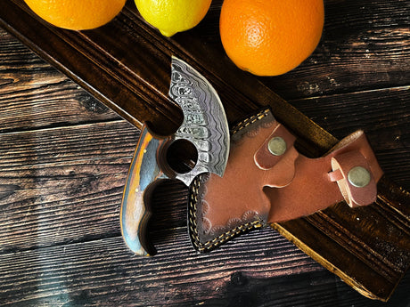 The Knife: Alaskan Ulu Style Blade, featuring a curved Damascus steel blade, ergonomic wooden handle, and leather sheath, rests on a wooden surface next to oranges and a lemon.