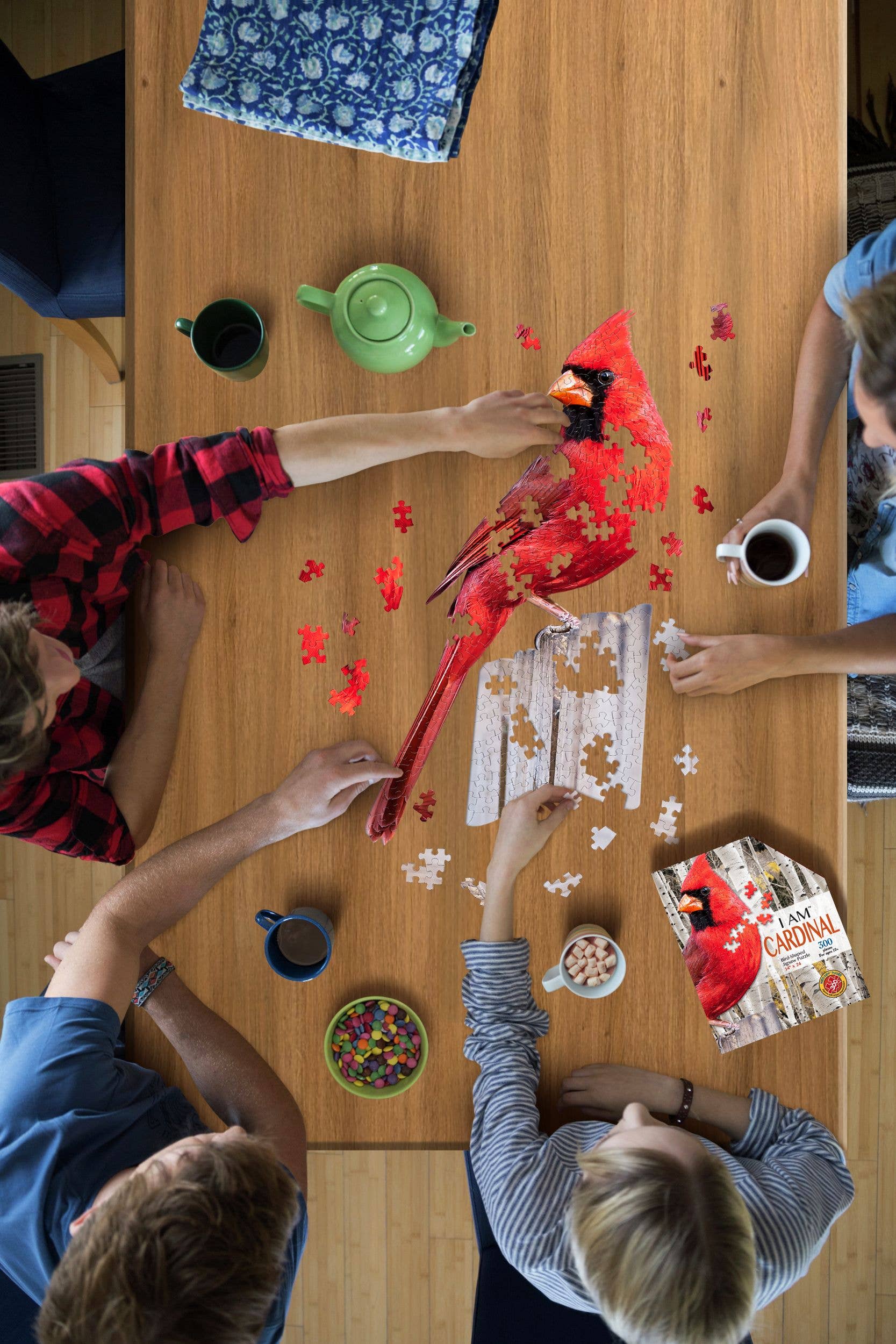 Four people gather around a wooden table, working on the I AM CARDINAL 300 piece educational puzzle while enjoying drinks and a bowl of candies.