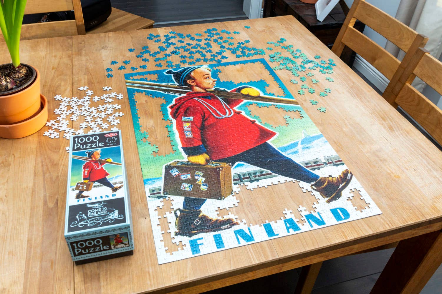 A partially completed Ski-Boy Finland 1000 pc Puzzle featuring a person in a red coat with a suitcase sits on the wooden table. Nearby, a vintage Ski-boy poster casually leans against a potted plant and the puzzle box.