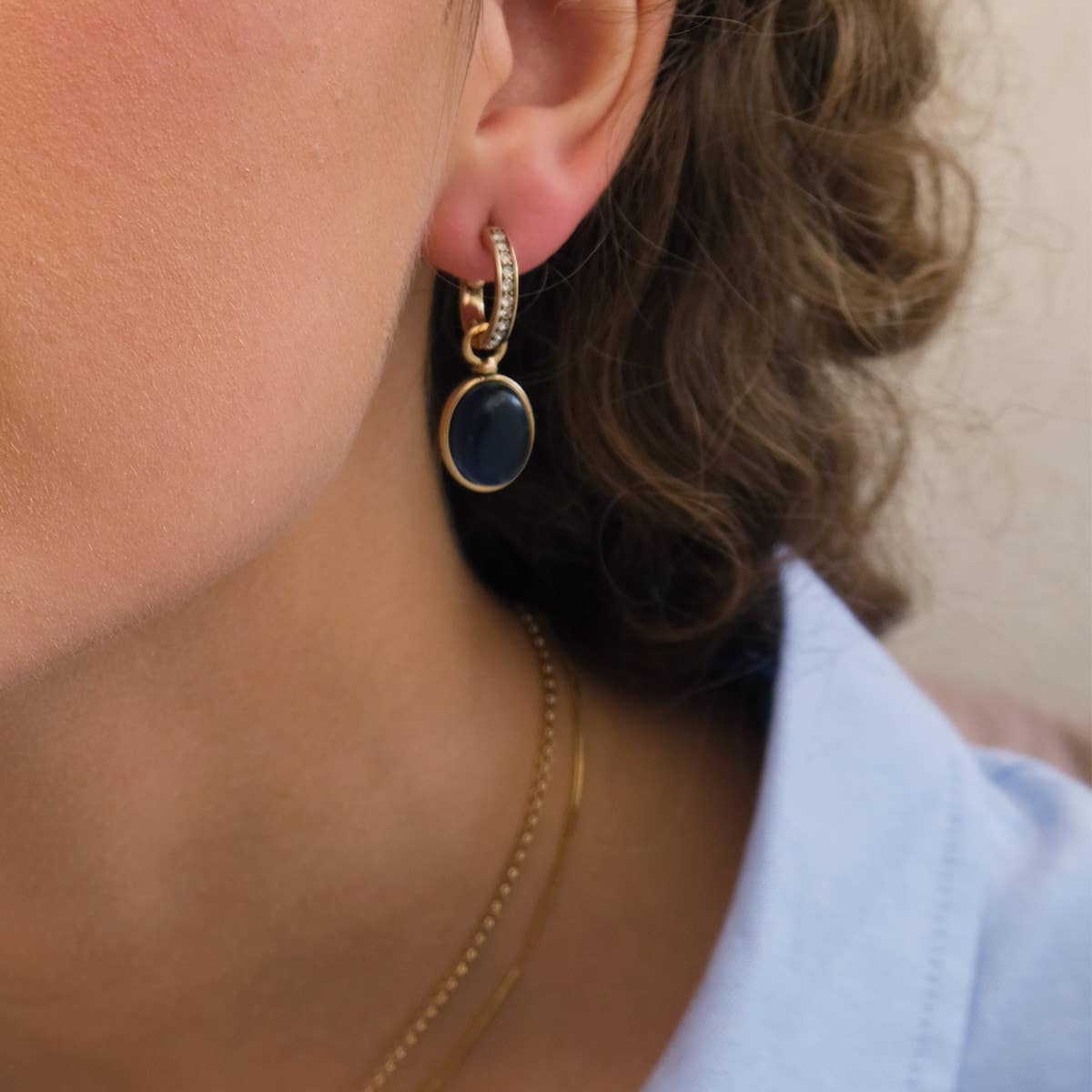 Wearing A&C Oslo Gold Plated Glass and Crystal Pendant Earrings with a dangling black oval charm and a light blue collared shirt, the person showcases elegance.