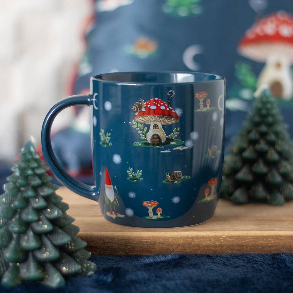 The Gnome Sweet Gnome Print Mug, featuring a whimsical mushroom house design reminiscent of a winter forest, sits on a wooden surface with small decorative tree figures surrounding it.