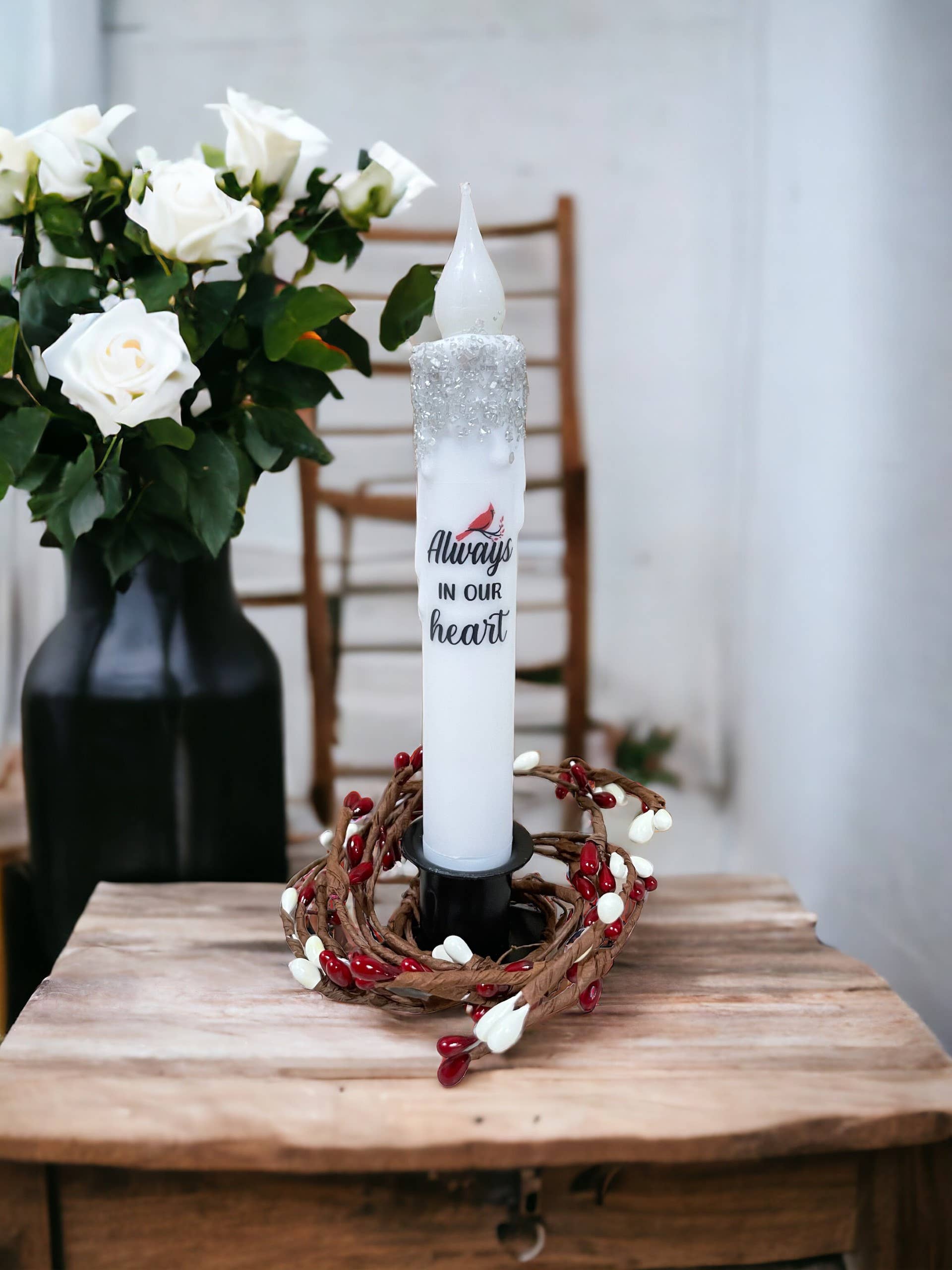 The Cardinal Memorial LED Timer Candle, inscribed with Always in our heart and adorned with a berry garland, features a flickering LED bulb. This flameless sympathy gift is set on a wooden table, complemented by a black vase of white roses in the background.