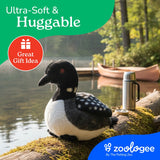 A soft, plush Earth Keeperz Loon Stuffed Animal by Faire - The Petting Zoo—crafted from recycled plastic water bottle fabric—rests on a mossy log near a lake, with a canoe, thermos, and trees in the background. Measuring 12″ long by 9″ high, this sustainable Plush: Loon features text reading “Ultra-Soft & Huggable” and “Great Gift Idea,” with the Zoologee logo at the bottom.