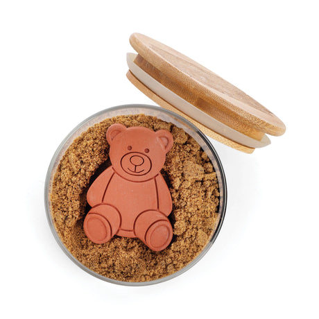A round glass jar filled with brown sugar holds a Sugar Saver: Terracotta Brown Sugar Bear Keeper & Saver, 2.5", shaped like a bear, with a wooden lid resting on the side.