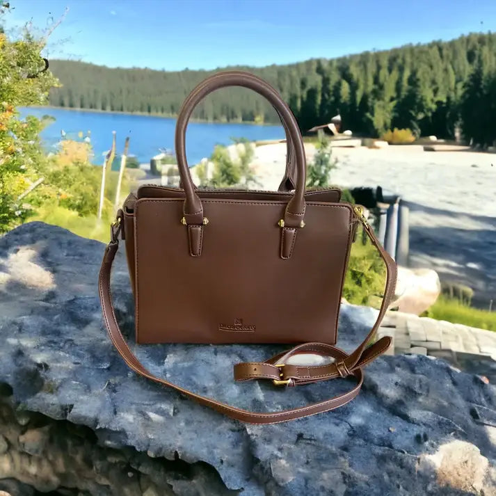 The Harris Tweed Double Handle Tote - Dark Brown, featuring double handles and a shoulder strap, is placed on a stone surface outdoors with a lake and pine trees in the background.